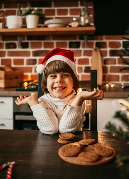 白いニットのセーターと赤いクリスマスの帽子の幸せな男の子がキッチンテーブルに座っています — ストック写真