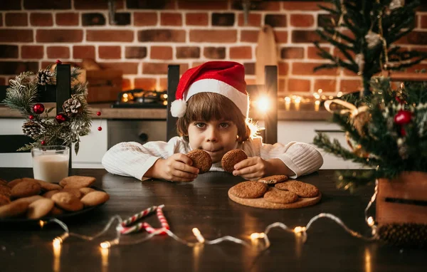 Beyaz örgü süveter ve kırmızı Noel şapkalı küçük bir çocuk mutfak masasında oturmuş yulaflı kurabiyeler yiyor.