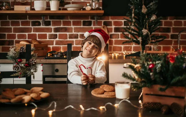 白いニットのセーターと赤いクリスマスの帽子の男の子が台所のテーブルに座ってキャンディー缶を持っています — ストック写真