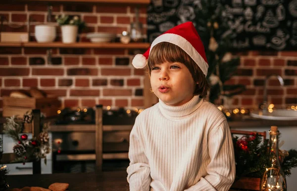 白いニットのセーターとキッチンのテーブルに座っている赤いクリスマスの帽子の小さな男の子の肖像画 — ストック写真