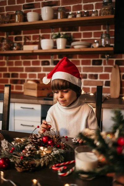 白いニットのセーターと赤いクリスマスの帽子の男の子がキッチンに座って自分の手でクリスマスの花輪を作っています — ストック写真