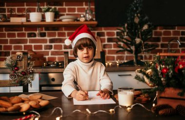 Beyaz süveter ve kırmızı Noel şapkalı küçük bir çocuk mutfak masasında oturuyor ve yeni yıl mektubu yazıyor.