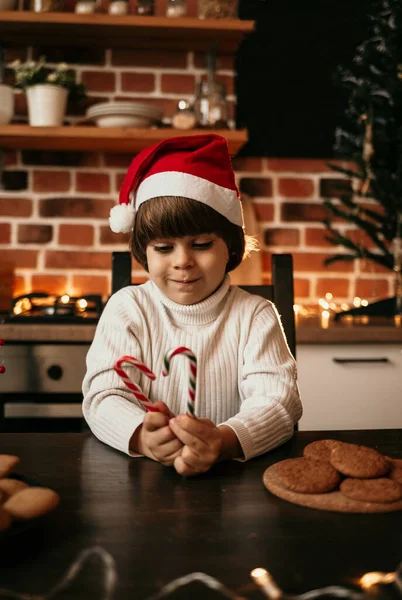白いセーターを着た可愛い男の子と赤いクリスマスの帽子がカラフルなキャンディーの杖でキッチンテーブルに座っています — ストック写真