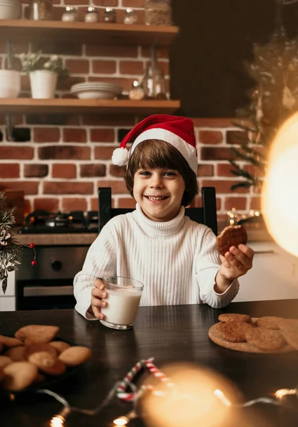 白いセーターに赤いクリスマスの帽子の幸せな男の子がミルクとオートミールクッキーとキッチンテーブルに座っています — ストック写真