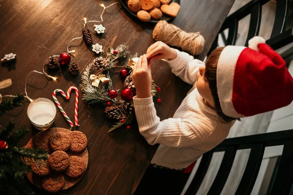 白いセーターと赤いクリスマスの帽子の男の子が台所のテーブルに座って自分の手でクリスマスの花輪を作っています — ストック写真