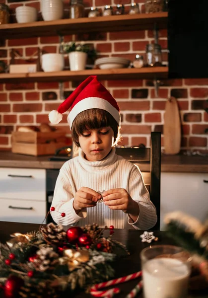 白いセーターを着た白人の男の子と赤いクリスマスの帽子が台所のテーブルに座って自分の手でクリスマスの花輪を作っています — ストック写真