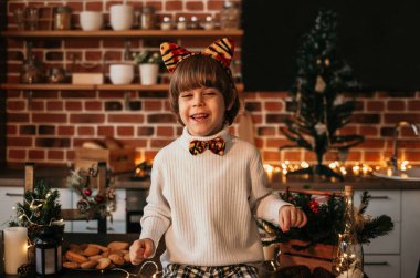 Küçük bir çocuk mutfak masasında beyaz bir kazak ve kaplan kulaklı bir saç bandıyla oturuyor.