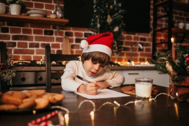 Yeni yıl çocuğu kırmızı bir yeni yıl şapkasıyla masada oturuyor ve bir kağıt parçasına resim çiziyor.