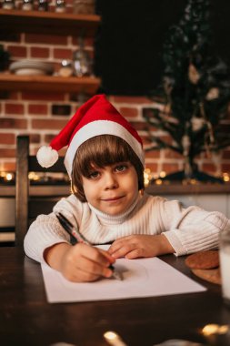 Yeni yıl çocuğu kırmızı bir yeni yıl şapkasıyla masada oturuyor ve bir kağıt parçasına yazı yazıyor.