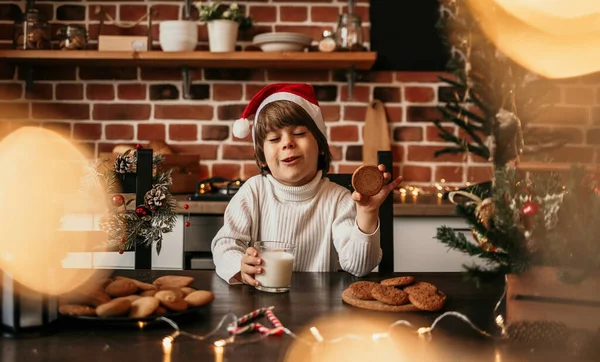 新年の子供は白いセーターのキッチンテーブルに座っていますミルクとクッキーの赤いクリスマスの帽子 — ストック写真