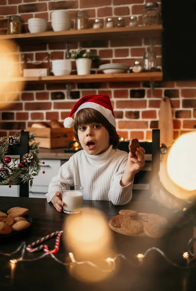 驚くべき白人の少年が白いセーターに赤いクリスマスの帽子にミルクとクッキーをつけてキッチンテーブルに座っています — ストック写真