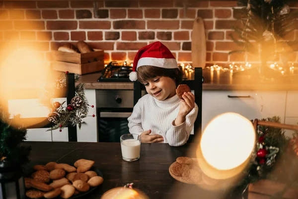 白人の男の子が白いセーターに赤いクリスマスの帽子にミルクとクッキーをつけて台所のテーブルに座っています — ストック写真