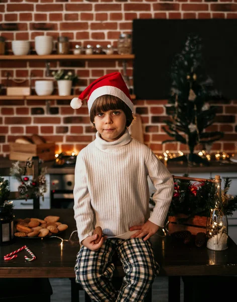 真剣な男の子が白いセーターと赤いクリスマスの帽子のキッチンテーブルの上に座っています — ストック写真