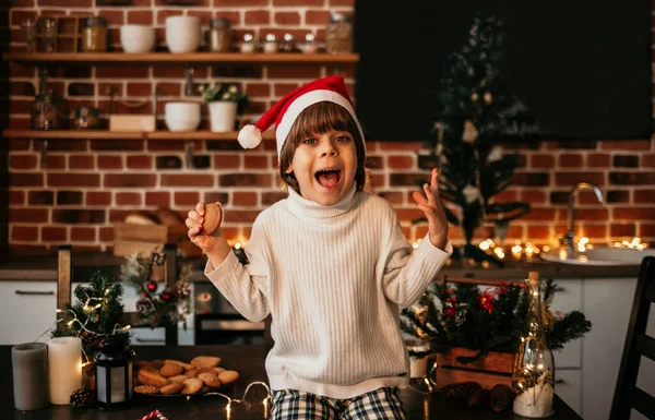 面白い小さな男の子は白いセーターと赤いクリスマスの帽子のキッチンテーブルの上に座っていますクッキーを手に — ストック写真