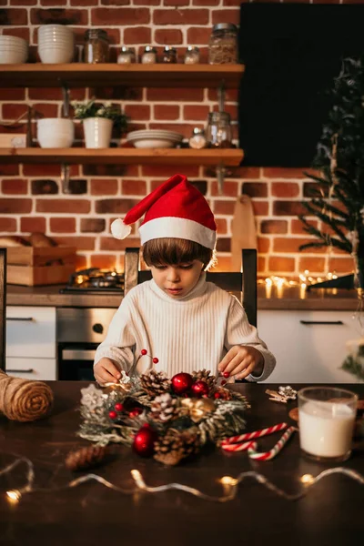 小さな男の子が赤いクリスマスの帽子のテーブルに座って自分の手でクリスマスの花輪を作っています — ストック写真