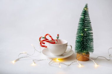 Bir fincan çay tabağı ve karamel kamışı ve beyaz arka planda küçük yapay bir Noel ağacı.