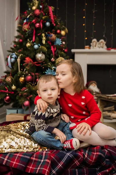 白人の弟と妹は部屋のクリスマスツリーの近くに座っていると抱擁 — ストック写真