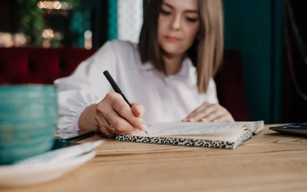 Beyaz bluzlu esmer bir iş kadınının bir kafedeki masada oturup not defterine not aldığı yakın plan fotoğrafı.