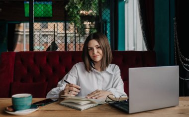 Güzel bir esmer iş kadını bir kafede dizüstü bilgisayarla oturuyor ve not defterine notlar yazıyor.