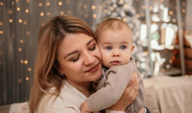 Sarışın bir annenin, bir bebeği Noel ağacının arka planında tutarken çekilmiş hassas bir fotoğrafı.