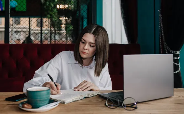 Bir iş kadını bir kafede dizüstü bilgisayarla oturuyor ve not defterine not alıyor.