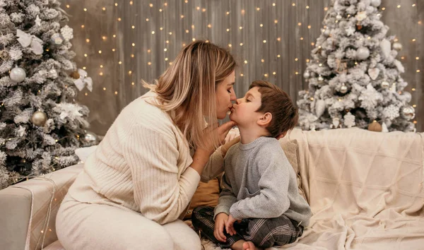 ブロンドのお母さんはソファに座って子供とキスしてるクリスマスツリーを背景に — ストック写真