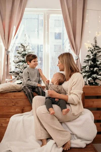 お母さんは子供たちと一緒に窓の近くの部屋に座っているクリスマスツリーを持つ息子 — ストック写真