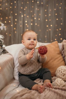Bir erkek bebek Noel ağacı olan bir odada elinde oyuncakla kanepede oturuyor.