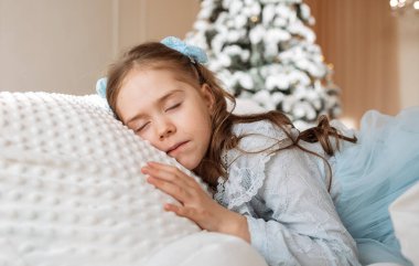 Şenlikli elbiseli küçük bir kız Noel ağacı olan bir odada beyaz bir yatakta uyuyor.