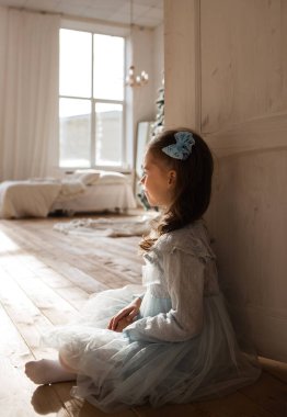 Mavi elbiseli küçük bir kız odanın kapısının yanında oturuyor.