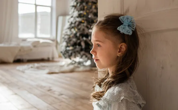 青いドレスを着た女の子がクリスマスツリーのある部屋のドアの近くに座って — ストック写真
