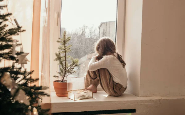Üzgün küçük bir kız Noel ağacının yanındaki bir odanın pencere eşiğinde oturuyor.