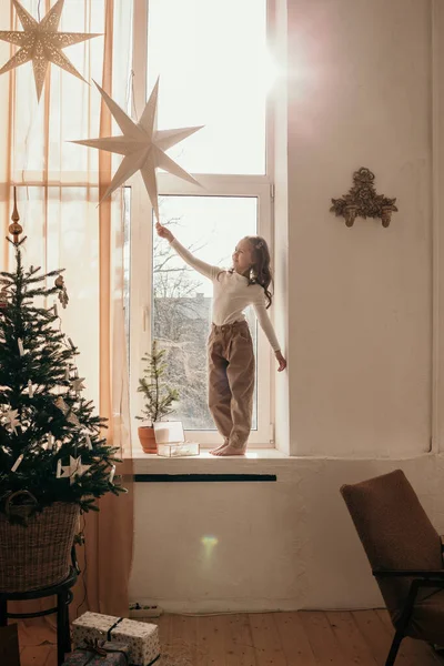 窓辺に小さな女の子が立って星のガーランドで遊んでいます — ストック写真