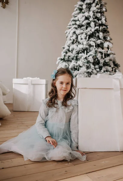 青いお祝いのドレスを着た女の子が部屋のクリスマスツリーの近くの巨大な贈り物の隣に座っています — ストック写真