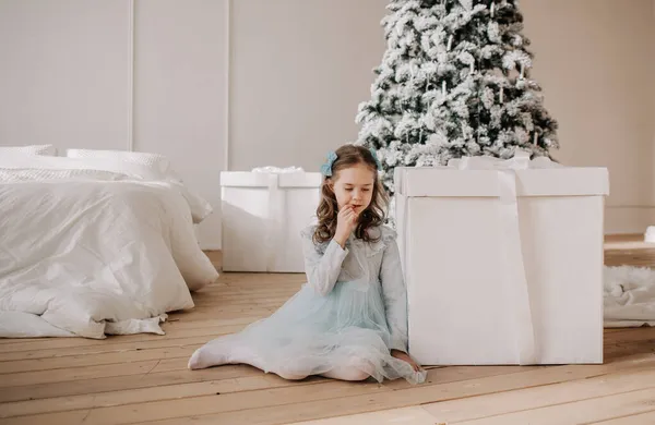 青いお祝いのドレスを着た思慮深い女の子が部屋のクリスマスツリーの近くの巨大な贈り物の隣に座っています — ストック写真