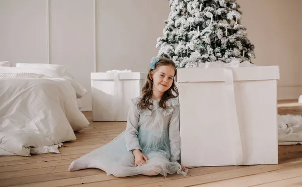 青いお祝いのドレスを着た笑顔の女の子が部屋のクリスマスツリーの近くの巨大な贈り物の隣に座っています — ストック写真