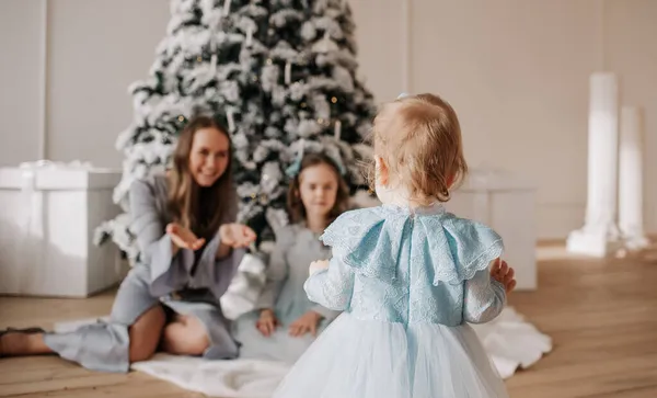 小さな娘が後ろに立ってクリスマスツリーの部屋で母親と妹に最初の一歩を踏み出す — ストック写真