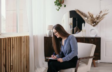 Gözlüklü esmer bir psikolog odada kağıtları olan bir sandalyede oturuyor ve not alıyor. Profesyonel psikolojik yardım.