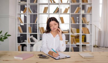 Bir iş kadını, kitaplarla dolu bir odada bir masaya oturur ve belgeleri doldurur.