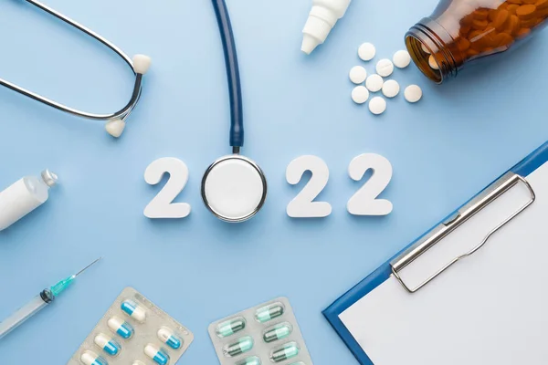2022 Happy New Year banner for health care and medical concept. Stethoscope with doctor order chart, prescription, pills, syringe, vaccine vial and white number 2022 on table blue background.