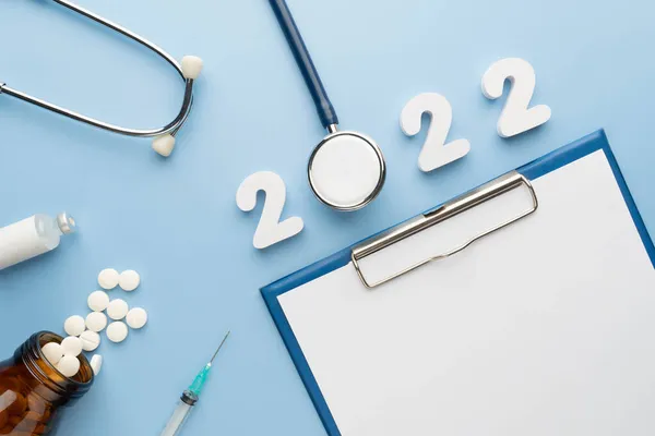 2022 Happy New Year banner for health care and medical concept. Stethoscope with doctor order chart, prescription, pills, syringe, vaccine vial and white number 2022 on table blue background.