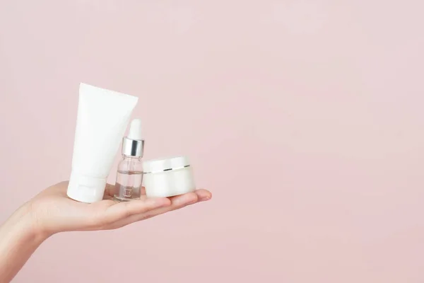 Skincare routine for healthy skin. Woman hand holding facial foam tube, serum bottle, moisturizing cream on pink background. Beauty and cosmetics concept.