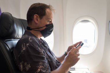 Uçakta telefonuna bakarken maske takan ve uzatan bir adam.