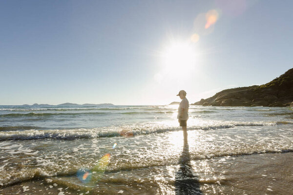 A caucasian boy child wearing a hat standing alone in the water at an australian beach with waves crashing around him