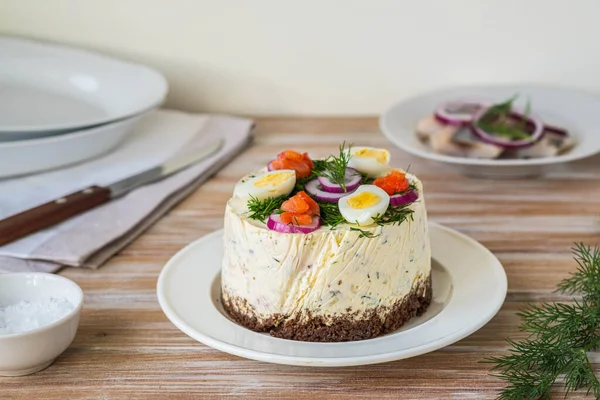 Snack, round Swedish salad cake with herring, potatoes, egg and sour cream mousse on black bread on a light plate on a wooden background. Christmas and Easter recipes. Swedish cuisine.