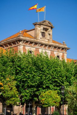 İspanya 'nın Barselona kentindeki tarihi binanın tepesinde dalgalanan bir direkte İspanyol ve Katalan bayrakları. Mavi gökyüzü ve yeşil ağaçlar parkta.