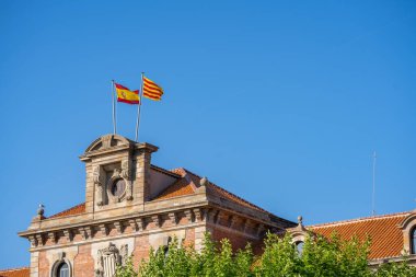 Spanish and Catalan flags on a pole waving on top of the historical building in Barcelona, Spain. Blue sky and green trees in a park.