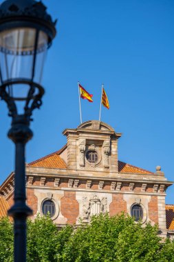 İspanya 'nın Barselona kentindeki tarihi binanın tepesinde dalgalanan bir direkte İspanyol ve Katalan bayrakları. Mavi gökyüzü ve yeşil ağaçlar parkta.