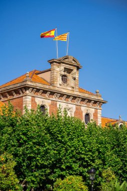 İspanya 'nın Barselona kentindeki Katalonya Parlamentosu' nun tepesinde İspanyol ve Katalan bayrakları dalgalanıyor. Mavi gökyüzü ve yeşil ağaçlar parkta.
