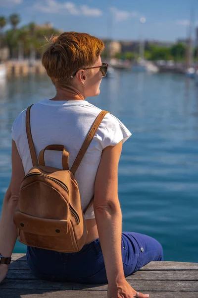 Young woman with backpack, seating in the port or harbor or marina with luxury yachts in Barcelona on the background. View from the back.